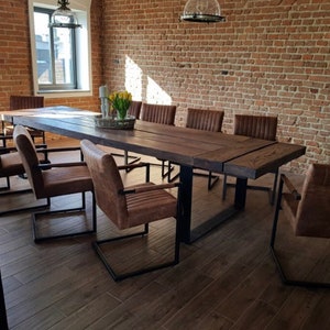 May include: A long wooden table with a black metal frame sits in a room with exposed brick walls. The table is surrounded by brown leather chairs with black metal frames.