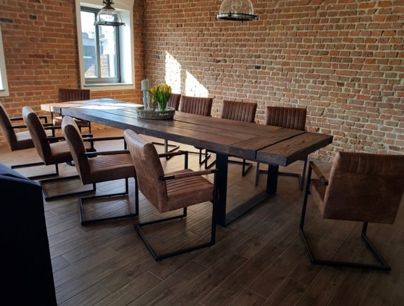 May include: A long wooden table with a black metal frame sits in a room with exposed brick walls. The table is surrounded by brown leather chairs with black metal frames.