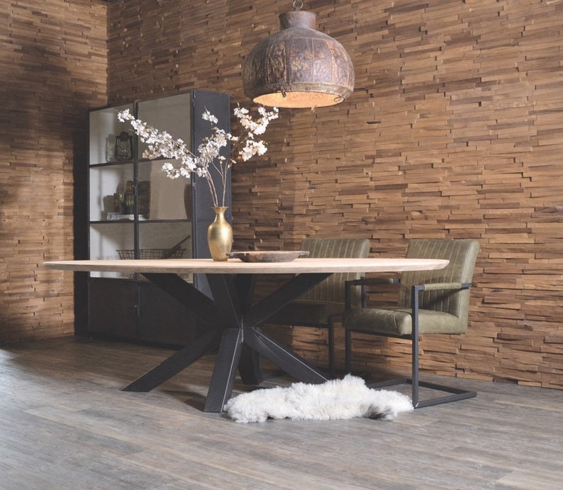 May include: A large oval wooden dining table with a black metal base and a starburst design. Two green upholstered chairs with black metal frames are placed at the table. A white faux fur rug is on the floor. The wall behind the table is covered in wood paneling.