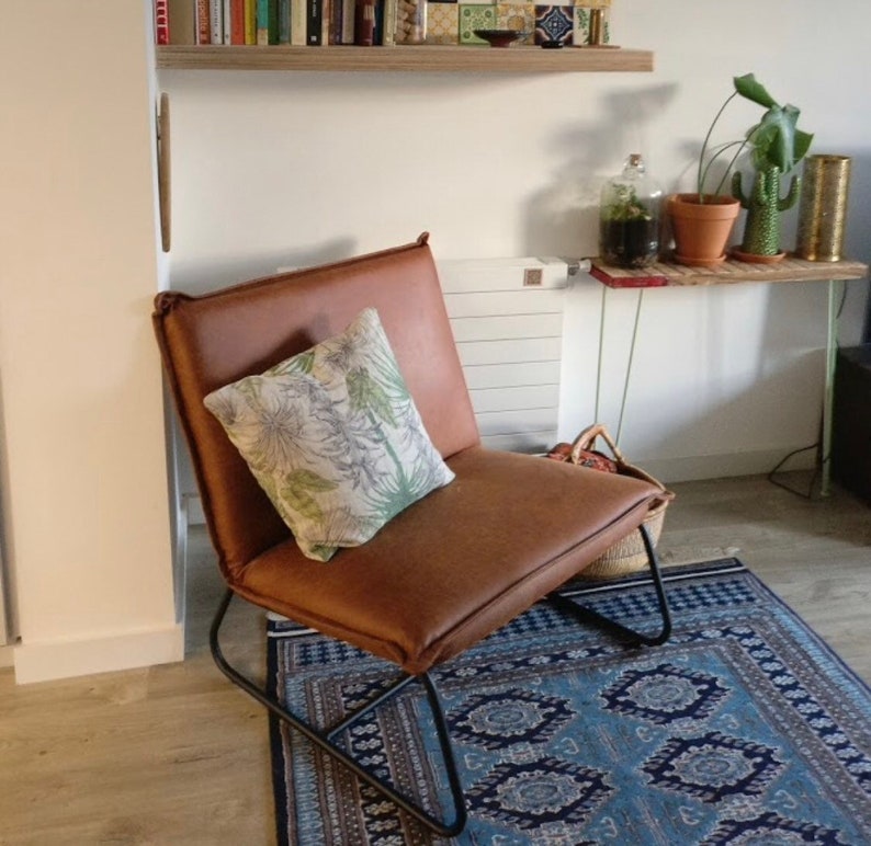 Puede incluir: Un sill&oacute;n de cuero marr&oacute;n con un marco de metal negro se encuentra sobre una alfombra de patr&oacute;n azul y blanco. Un coj&iacute;n blanco con estampado floral verde est&aacute; en el sill&oacute;n.