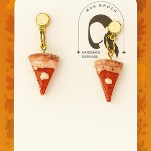 Può includere: Orecchini fatti a mano con ciondoli a forma di fetta di torta in miniatura. Ogni ciondolo è una fetta triangolare con ripieno rosso, crosta marrone e una macchia bianca. Gli orecchini pendono da perni e catene color oro. La carta dice "RVA BRUSH HANDMADE EARRINGS."