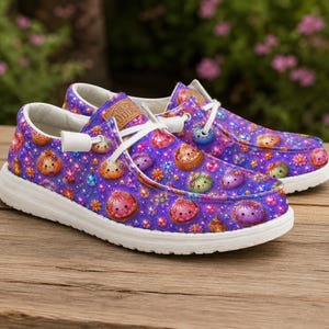 K&ouml;nnte beinhalten: Ein Paar Slipper mit einem leuchtend violetten Hintergrund, der ein wiederkehrendes Muster aus kleinen, bunten, runden Kreaturen und funkelnden Sternen zeigt. Die Schuhe haben wei&szlig;e Schn&uuml;rsenkel, wei&szlig;e Sohlen und einen hellbraunen Aufn&auml;her.