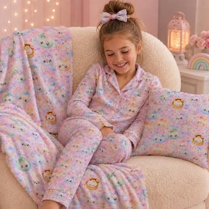 K&ouml;nnte beinhalten: Ein lang&auml;rmeliges Schlafanzug-Set mit Knopfleiste, ein passendes quadratisches Zierkissen und eine weiche Decke, alle mit einem wiederkehrenden Muster aus pastellfarbenen Kawaii-Kn&ouml;deln mit l&auml;chelnden Gesichtern auf hellviolettem Grund.