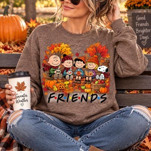 Peut inclure: Un sweat-shirt marron à col rond orné d'un motif représentant les personnages de Peanuts assis sur un banc entourés de feuilles d'automne, de citrouilles et d'un panier de pommes. Le texte "FRIENDS" est imprimé sous les personnages et une tasse blanche indique "Sweater Weather".