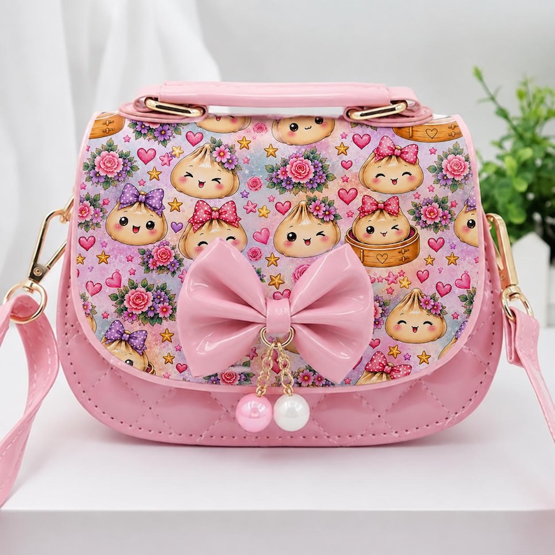 Puede incluir: Un bolso bandolera rosa con acabado brillante y panel inferior acolchado. La solapa muestra un patr&oacute;n de dumplings de dibujos animados, flores rosas y corazones peque&ntilde;os. Un lazo rosa brillante est&aacute; centrado al frente con dos perlas colgantes.