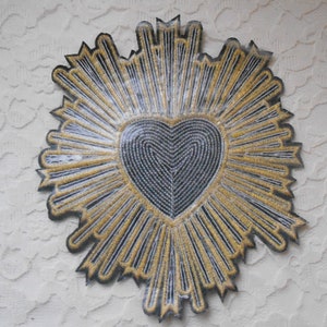 May include: A black felt heart with gold embroidery, surrounded by a radiating sunburst design. The heart is outlined in black and filled with a swirling pattern.