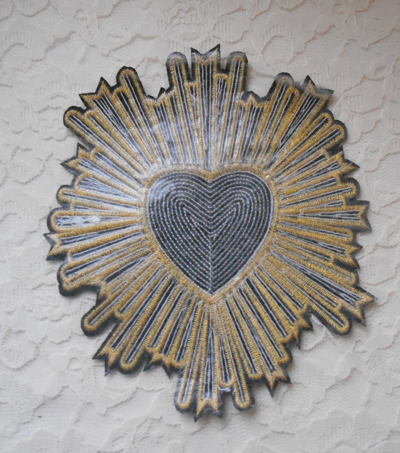 May include: A black felt heart with gold embroidery, surrounded by a radiating sunburst design. The heart is outlined in black and filled with a swirling pattern.