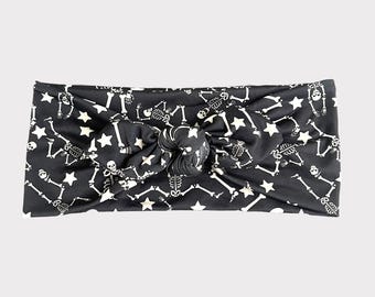 Op de afbeelding: Zwart-wit gepatroneerd hoofdband met een skelet- en sterrendesign. Het hoofdband is in een knoop geknoopt.