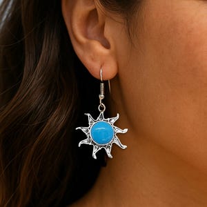 May include: Silver-toned sunburst earrings with a bright turquoise center stone. The earrings are dangling from a silver hook. The background is a blurred view of the ocean and sky.