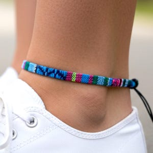 May include: A colorful woven ankle bracelet with blue, green, pink, and purple stripes is worn above a white sneaker. The bracelet is secured with a black string. The image shows a close-up of the ankle and shoe.