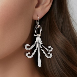 May include: Silver chandelier earrings with a unique design. The earrings feature a curved top with multiple dangling, stylized, curved elements. The earrings are worn by a person with brown hair and eyes, and a neutral background.