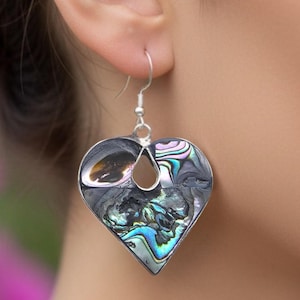 May include: Heart-shaped earrings with iridescent abalone shell inlays. The earrings have a silver-tone metal frame and a teardrop cutout at the top. The abalone shell displays shades of blue, green, and grey.