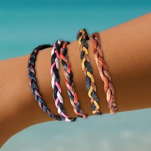 May include: A close-up of a person's wrist wearing five braided bracelets. The bracelets are in various colours, including black, white, pink, orange, and yellow.