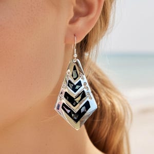 May include: Silver-toned earrings with a geometric, layered design. The earrings feature a chevron pattern with dark blue and iridescent accents. The earrings are dangling, with a hook closure. The background is a beach scene.