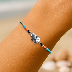May include: A beaded bracelet with a silver elephant charm. The bracelet features turquoise, red, and black beads. The elephant charm is detailed with etched patterns. The background shows a beach scene with a woven bag.