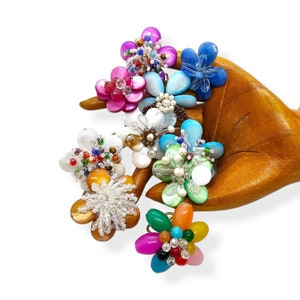 May include: A collection of colorful beaded flower rings. The rings are made with various beads, including pearls, glass beads, and shell beads. The rings are arranged in a hand, with the flowers facing outwards.