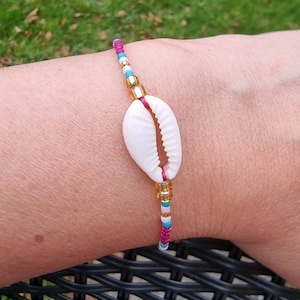 May include: A bracelet featuring a central white cowrie shell strung on a vibrant pink cord. The design is accented with small, multi-coloured glass beads in shades of turquoise, white, and metallic gold, arranged symmetrically on either side.