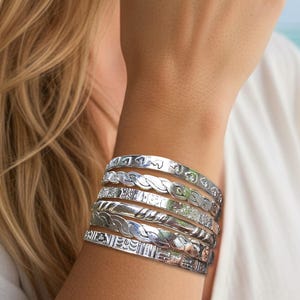 May include: A collection of silver bangles with various engraved designs, including floral and geometric patterns. The bracelets are stacked on a wrist, showcasing their intricate details and reflective surfaces. The background features a beach scene with palm trees.