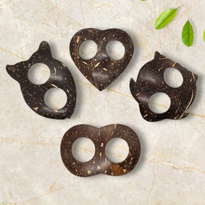 May include: Four dark brown coconut shell finger holders in various shapes: a cat, a heart, an owl, and a figure eight. Each has two circular finger holes. The natural wood grain is visible throughout the pieces.