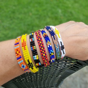 May include: A set of colorful beaded bracelets with a mix of yellow, red, blue, black, and white beads. The bracelets are stacked on a person's wrist.