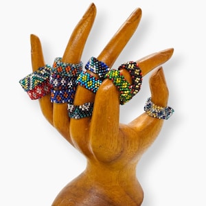 Könnte beinhalten: Ein hölzerner Hand-Ringständer mit sechs bunten Perlenringen. Die Ringe sind in verschiedenen Farben gefertigt, darunter Blau, Grün, Rot, Gelb und Schwarz.