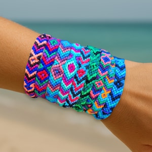 May include: A selection of vibrant friendship bracelets, each with a unique woven design, stacked on a wrist. The bracelets display a range of colours including blue, pink, green, and purple. The background shows a blurred ocean view.