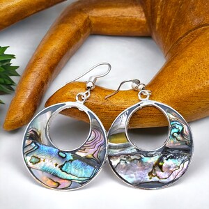 May include: Silver hoop earrings with iridescent abalone shell inlays. The earrings have a circular design with a silver frame and a hook closure. The abalone shell shows blue, green, and purple colours.