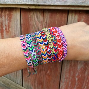 May include: A stack of colorful friendship bracelets with geometric patterns in shades of pink, green, blue, orange, and yellow. The bracelets are woven with a chevron pattern and are tied together on a person's wrist.