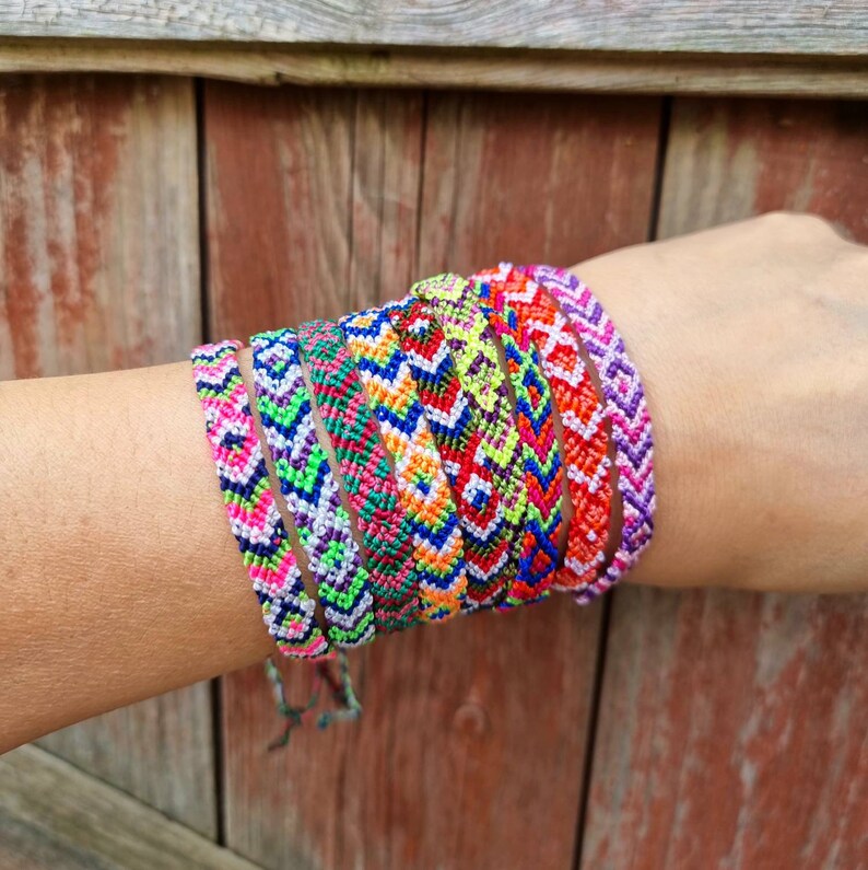 May include: A stack of colorful friendship bracelets with geometric patterns in shades of pink, green, blue, orange, and yellow. The bracelets are woven with a chevron pattern and are tied together on a person's wrist.