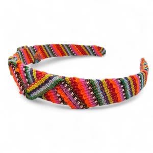 May include: A brightly coloured knotted headband with a striped pattern. The headband features orange, pink, green, blue, and yellow stripes. The fabric appears woven, giving a textured appearance. The headband is designed to be worn around the head.