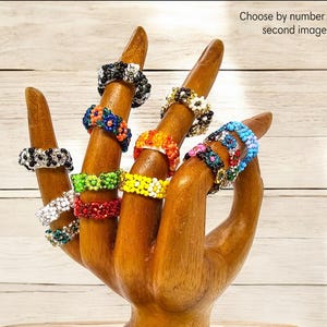 May include: A wooden hand model displaying a variety of colorful beaded rings. The rings are made with small beads and feature different floral patterns.