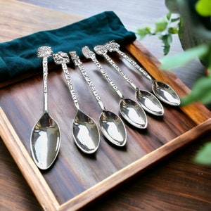 May include: Set of five silver-colored spoons with detailed handles featuring carved faces and symbols. The spoons are arranged on a wooden tray with a dark green cloth, showcasing a vintage or antique aesthetic.