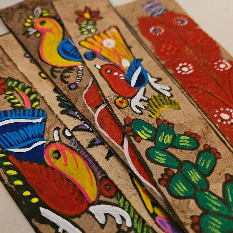 May include: A collection of handmade bookmarks with colorful, hand-painted designs. Each bookmark features a unique illustration of birds, flowers, and cacti in vibrant hues of yellow, red, blue, and green on a textured, tan background.