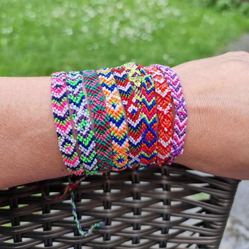 May include: A stack of colorful friendship bracelets with geometric patterns in shades of pink, purple, blue, green, orange, red, and white.