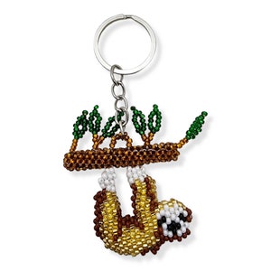 May include: A beaded sloth keychain hanging from a silver key ring. The sloth is brown and yellow with white accents and is hanging from a brown branch with green leaves.