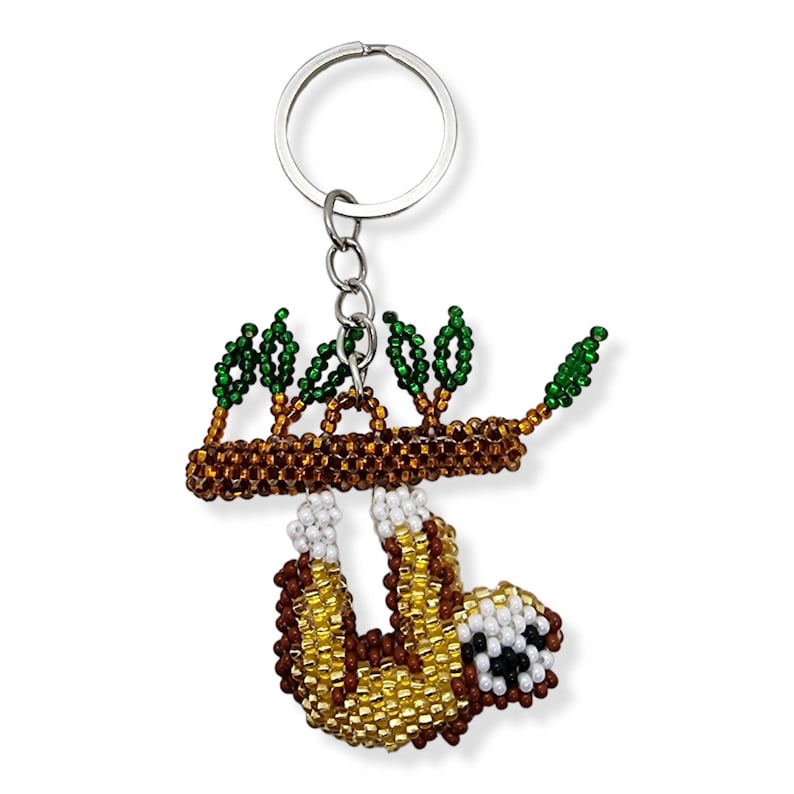 May include: A beaded sloth keychain hanging from a silver key ring. The sloth is brown and yellow with white accents and is hanging from a brown branch with green leaves.