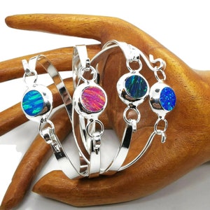 May include: Four silver bracelets with colourful opal stones. Each bracelet features a round opal stone in shades of blue, pink, and green, set in a silver bezel. The bracelets are displayed on a wooden hand.