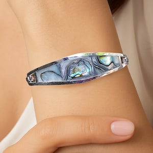 Peut inclure: Un bracelet argenté avec un incrustation en nacre d'ormeau. La coquille présente des reflets bleus, verts et violets irisés, sertis dans un cadre argenté. Le bracelet est porté sur un poignet, mettant en valeur son design élégant.