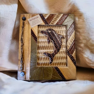 May include: A brown leather notebook with a dolphin design on the front cover. The notebook is made from natural leaf and wood. The text on the label reads "This notebook is handcrafted from natural leaf and wood."