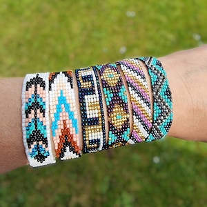 May include: A selection of beaded bracelets in diverse colours and patterns. The bracelets display designs in white, black, turquoise, pink, gold, and navy blue. The bracelets are worn on a wrist against a blurred green backdrop.