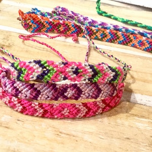 May include: Three colorful friendship bracelets with intricate woven patterns in pink, purple, green, white, and blue.