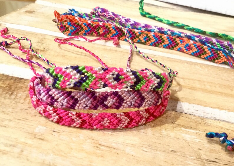 May include: Three colorful friendship bracelets with intricate woven patterns in pink, purple, green, white, and blue.