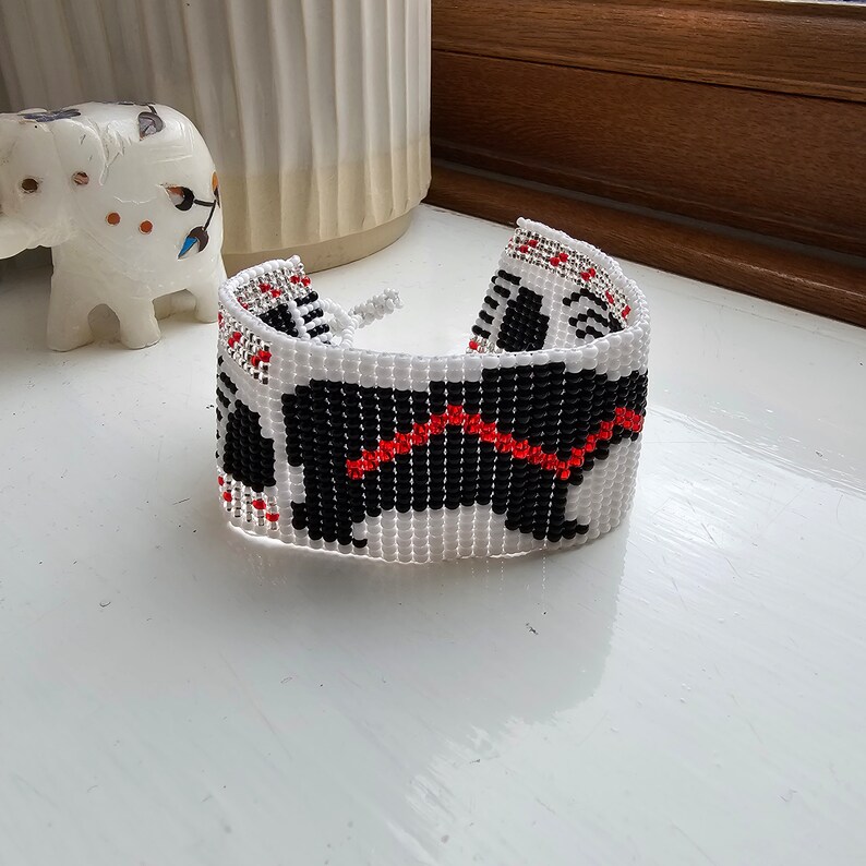 May include: White beaded bracelet with a black and red design of a dog. The dog is outlined in black and has a red line across its back.