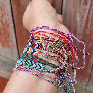 May include: A stack of colorful friendship bracelets made with woven string. The bracelets are tied together and feature a variety of patterns and colors, including red, blue, green, yellow, and purple.