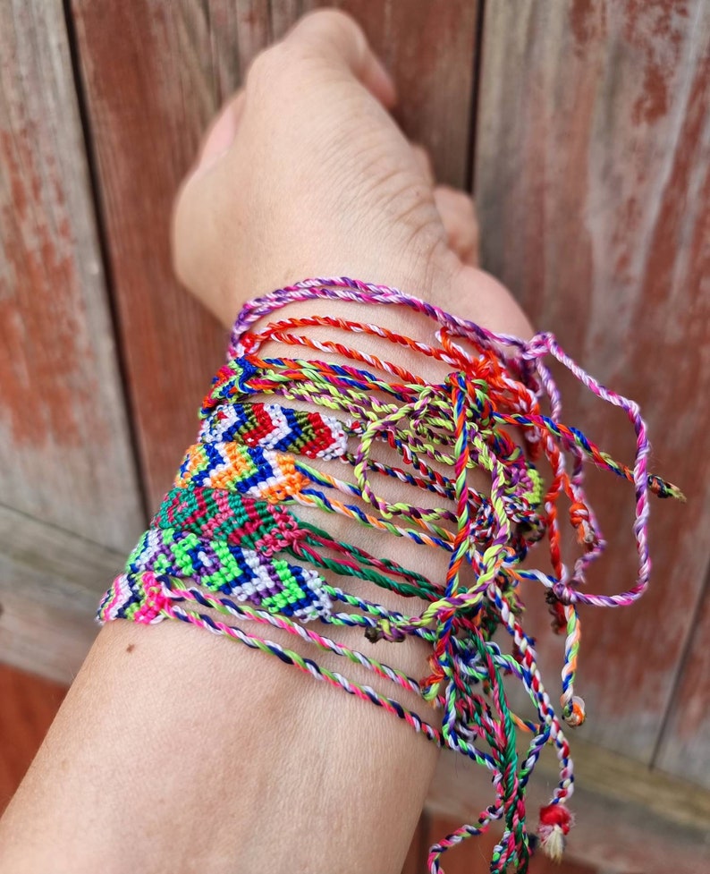 May include: A stack of colorful friendship bracelets made with woven string. The bracelets are tied together and feature a variety of patterns and colors, including red, blue, green, yellow, and purple.