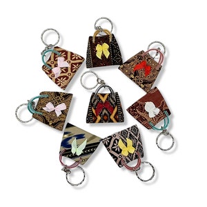 May include: A collection of fabric keychains shaped like handbags, each with a unique patterned design and a small bow. The keychains feature silver-colored metal rings and handles, and are arranged in a circular pattern against a white background.