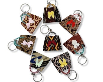 May include: A collection of fabric keychains shaped like handbags, each with a unique patterned design and a small bow. The keychains feature silver-colored metal rings and handles, and are arranged in a circular pattern against a white background.