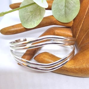 May include: Two silver-coloured, wavy-design bracelets. The bracelets are displayed on a wooden hand-shaped display, with a white background and a green and white floral accent.