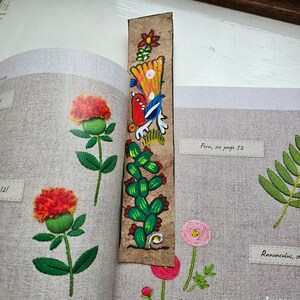 May include: A handmade bookmark with a colorful, painted design featuring a bird, cactus, and flowers. The bookmark is made of a textured, brown material and is placed in a book with embroidered floral illustrations.