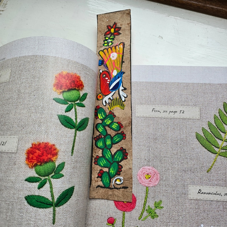 May include: A handmade bookmark with a colorful, painted design featuring a bird, cactus, and flowers. The bookmark is made of a textured, brown material and is placed in a book with embroidered floral illustrations.